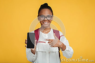 Satisfied smart adolescent black lady pupil in glasses with backpack shows finger at smartphone with blank screen Stock Photo