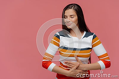 Satisfied woman keeps hands on belly, feels replete after delicious supper, has full stomach, long hair, appealing appearance, Stock Photo