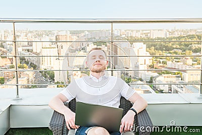Satisfied with work done. Young man with laptop keeping eyes closed, relaxing and dreaming while sitting on the terrace with Urban Stock Photo