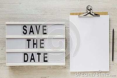 `Save the date` words on a modern board, clipboard with blank sheet of paper on a white wooden surface, top view. Overhead, from Stock Photo