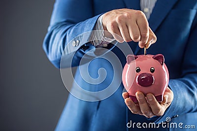 Saving money. Businesman holding pink piggy and putting coin into piggy bank Stock Photo