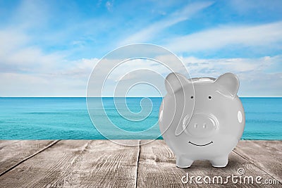Saving money for summer vacation. Piggy bank on wooden surface near sea, space for text Stock Photo
