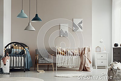 Scandi baby bedroom interior with an armchair between twin beds, standing against a beige wall with geometric paintings. Real Stock Photo