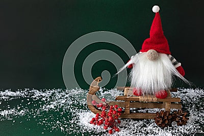 Scandinavian gnome with holly berry and pine sitting on wooden sledge on dark green background Stock Photo