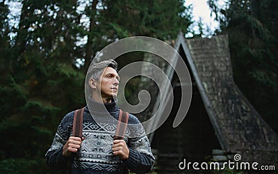 Scandinavian style portrait man in ethnic knitted sweater standing next to old wooden cabin in forest Stock Photo