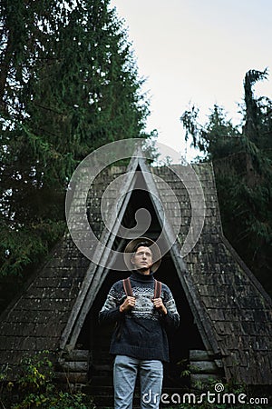 Scandinavian style vertical image man in ethnic sweater and hat standing by old wooden cabin in forest Stock Photo