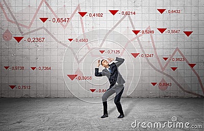 A scared businessman cowers near a concrete wall with red stock market indexes and falling statistic lines. Stock Photo