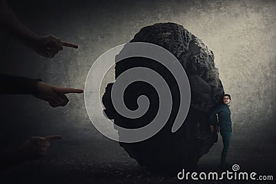 Scared businessman hiding behind a huge boulder, feels discomfort as people hands pointing to him blaming as guilty. High tension Stock Photo