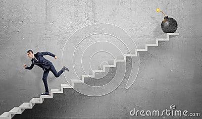 A scared businessman running down grey concrete stairs away from a large iron bomb with a lit fuse. Stock Photo