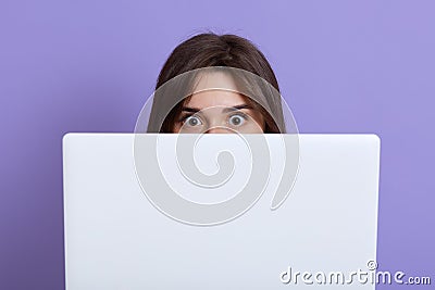 Scared dark haired female hiding behind laptop and looking at camera with eyes full of fear, being afraids of something, wants Stock Photo