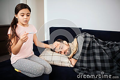 Scared small girl sitting besides sick father. She look at thermometer and wonder. Young man sleep. He sick and has Stock Photo