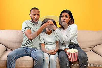 Scary moment in horror movie. African american parents closing daughter's eyes while watching TV, sitting on sofa Stock Photo