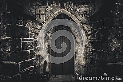 Scary pointy wooden door in an old and wet stone wall building with cross, skull and bones at both sides in black and white. Stock Photo