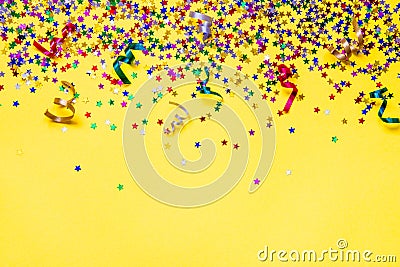 Scattered star shaped colorful glittering confetti and ribbons over yellow background. Copy space. Stock Photo