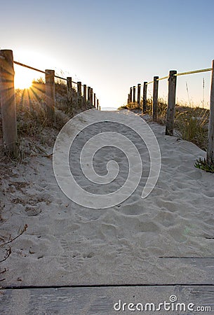 Scenic sunset on beach with wooden fence. Entrance to beach in evening sunlight. Wooden columns and path on sand. Stock Photo