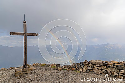 A scenics view of a mountain summit cross with a majestic double rainbow in the background under a stormy and rainy weather Stock Photo