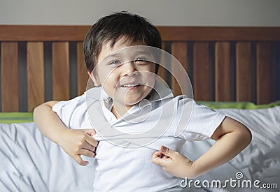 School boy sitting in bed and try to wearing his cloth with smiling face,Cute kid boy getting dressed and get ready for school, Stock Photo