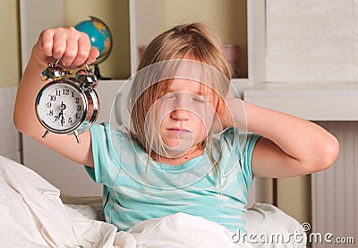 School girl hates waking up early in the morning. Blond girl holding alarm clock , yawning in bed covers ear with hand Stock Photo