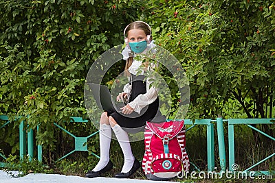 School girl in a medical mask, headphones engaged in a park on a laptop. Back to school. Virus protection for schools. Stock Photo