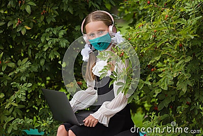 School girl in a medical mask, headphones engaged in a park on a laptop. Back to school. Virus protection for schools. Stock Photo