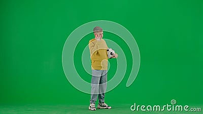 Portrait of kid boy on chroma key green screen. Schoolboy in jeans holding football ball and talking on smartphone. Full Stock Photo