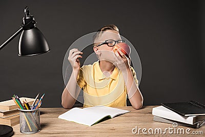 schoolboy in eyeglass eating apple and doing homework at table with lamp books colour pencils and textbook on grey Stock Photo