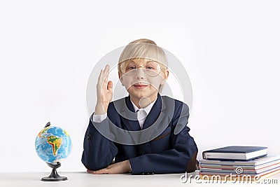 Schoolboy in large glasses sits at table with textbooks with his hand raised. Excellent student. Boy knows the answer Stock Photo