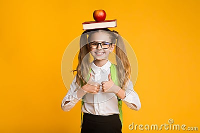 Schoolgirl Gesturing Thumbs-Up Holding Book And Apple On Head Stock Photo