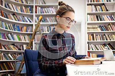 Schoolgirl in a plaid shirt leafing through a book while sitting in a library room. Young woman came to the library to prepare for Stock Photo