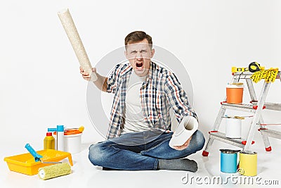 Screaming aggressive angry young man with rolls of wallpaper, sitting on floor with instruments for renovation apartment Stock Photo