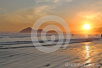 Sea with small waves, sand on the beach, mountains in the background, clouds from the sky, the sun sets, reflections in water and Stock Photo
