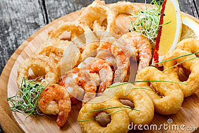 Seafood platter with deep fried squid rings, shrimp and onion rings decorated with lemon on cutting board on wooden background. Stock Photo