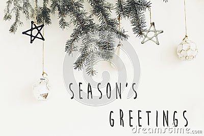 Season`s greetings text sign on stylish christmas branches with glass modern ornaments hanging on white wall. Creative christmas Stock Photo
