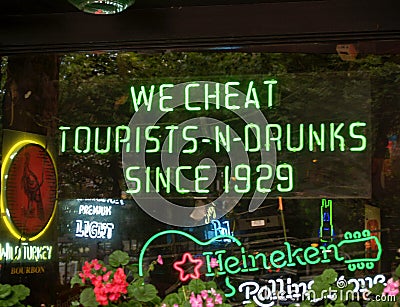 Bright green neon window sign photographed from street stating We Cheat Tourists-n-Drunks Since 1929 Editorial Stock Photo