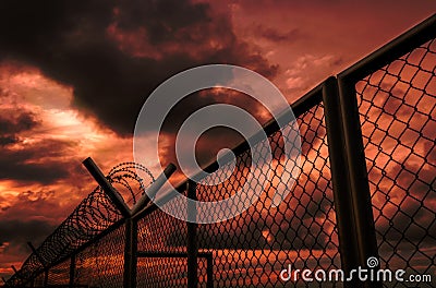 Security fence of military zone or private area fence with red sky and dark clouds. Barbed wire security fence with stormy sky. Stock Photo