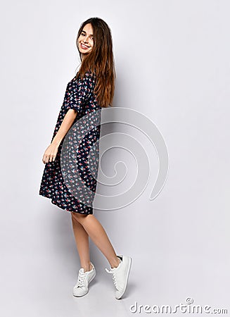 Brunette model in black dress with floral print, white sneakers. Smiling, touching her face, posing on white. Full length Stock Photo