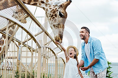 selective focus of cheerful man and kid with closed eyes feeding giraffe in zoo. Stock Photo