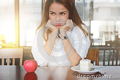 Selective focus on face of frustrated stressed Asian woman choosing between apple or unhealthy cake on the desk with sunshine effe Stock Photo