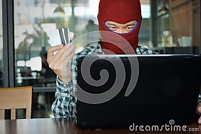 Selective focus on hands of Masked hacker wearing a balaclava holding credit card between stealing data from laptop. Internet crim Stock Photo