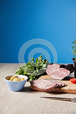 Focus of tasty ham on cutting board with knife, parsley, olives on wooden table on blue background Stock Photo