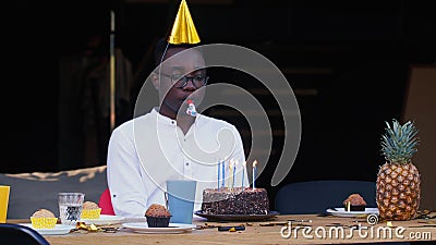 Self isolation. Sad lonely African young handsome man blows whistle alone celebrating his own birthday with cake at home Stock Photo