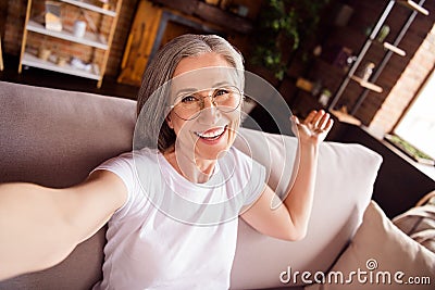 Self-portrait of attractive cheerful grey-haired woman sitting on divan welcoming you at loft industrial home house flat Stock Photo