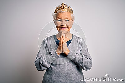 Senior beautiful grey-haired woman wearing golden queen crown over white background praying with hands together asking for Stock Photo