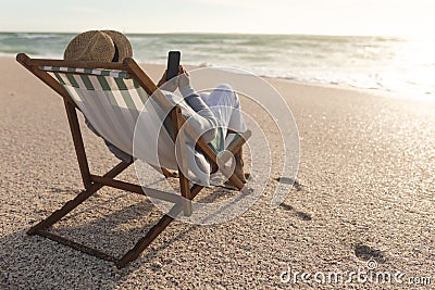 Senior biracial woman sitting on chair using mobile phone while relaxing at beach during sunset Stock Photo