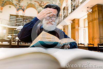 Senior concentrated man with white beard, touching his eyeglasses, sitting at the table and thinking about the book he Stock Photo