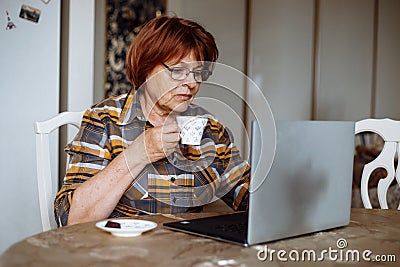 Senior concentrated woman sitting at table on white chair, holding cup of coffee, looking at laptop, working from home. Stock Photo