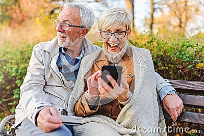 Senior couple sitting in public park using smart device to shop online Stock Photo