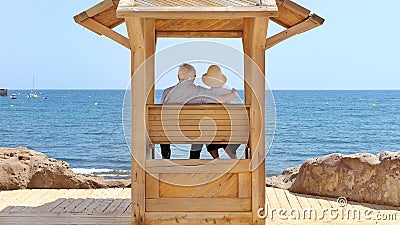 Senior couple, woman with hat and man with white silver hair sitting under a wooden canopy and looking at the ocean Stock Photo