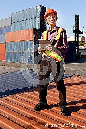 A senior elderly Asian worker engineer wearing safety vest and helmet standing and holding digital tablet at shipping cargo Stock Photo