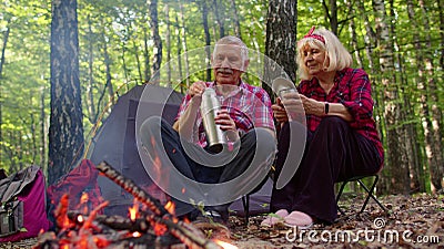 Senior elderly grandmother grandfather pouring drink from thermos drinking tea over campfire in wood Stock Photo